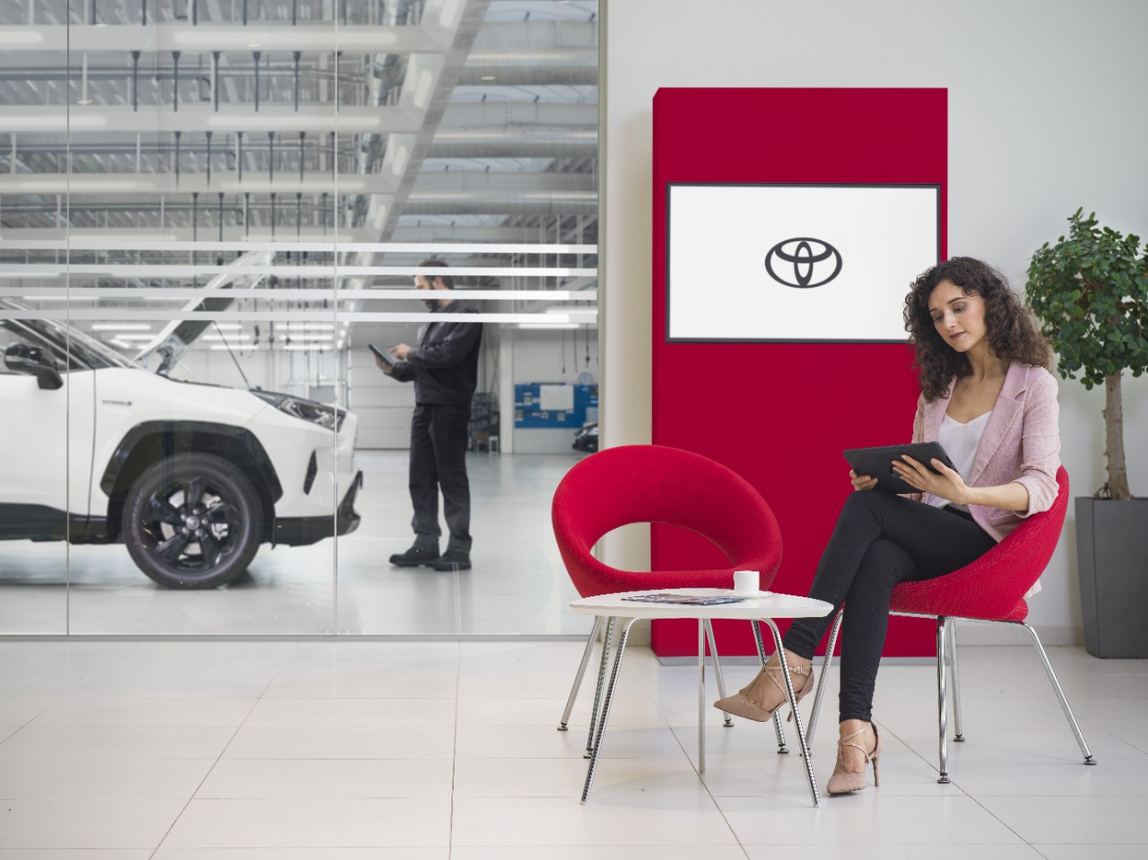 A women sits in a Toyota Service waiting area. She looks at a tablet, with a Toyota logo displayed on a tv screen behind her.